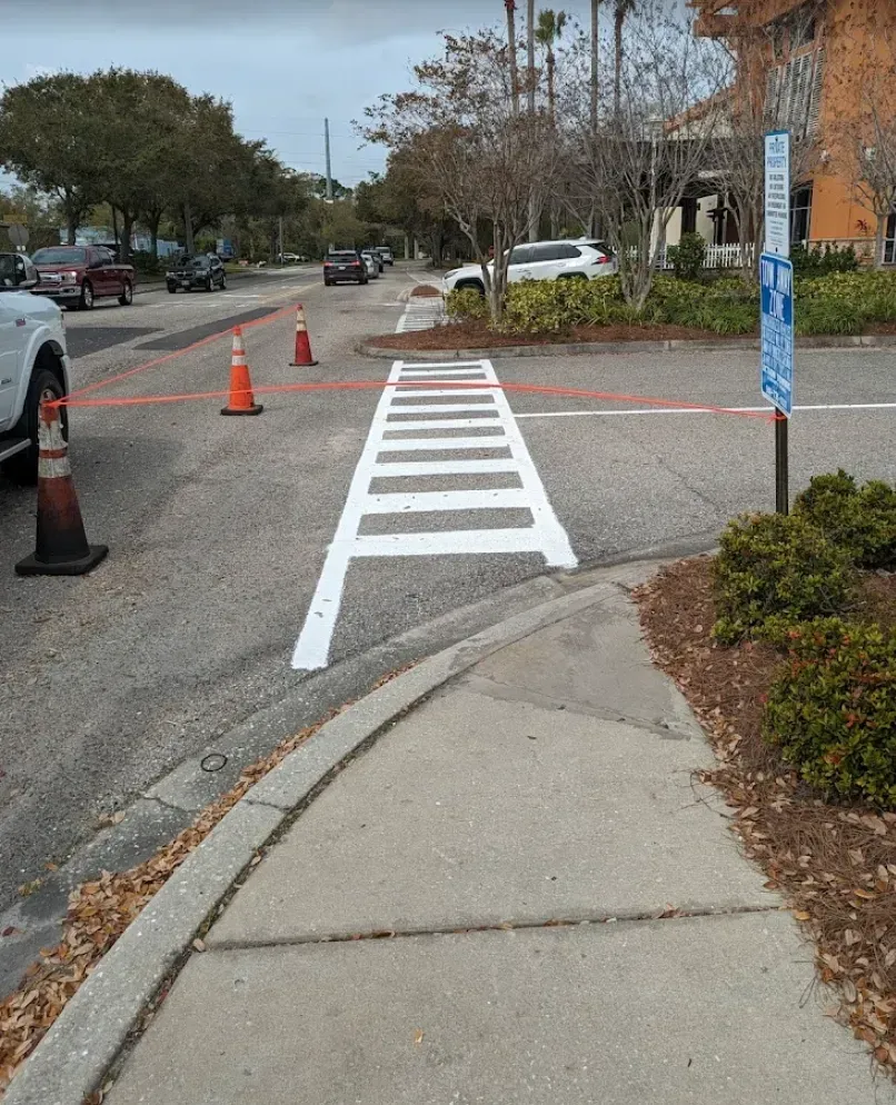 Temporary white crosswalk markings on a sidewalk, with orange cones blocking a parking lot entrance.