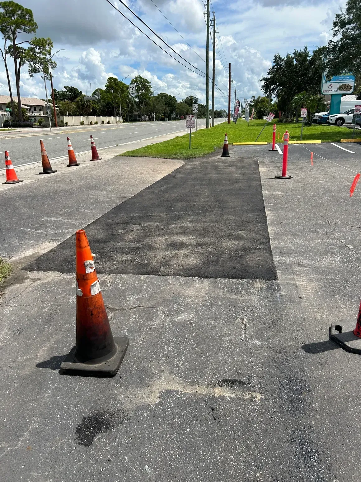 Roadwork lane with fresh asphalt patch, orange cones, and utility poles on a sunny street