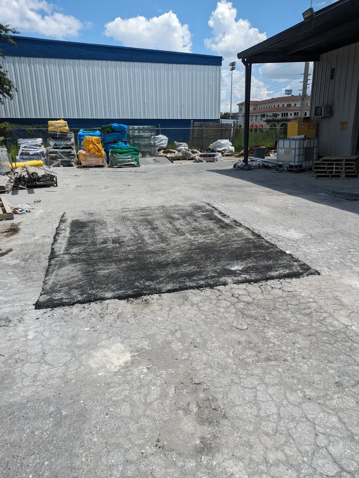 Outdoor industrial yard with a large black asphalt patch on cracked pavement, pallets and materials in the background