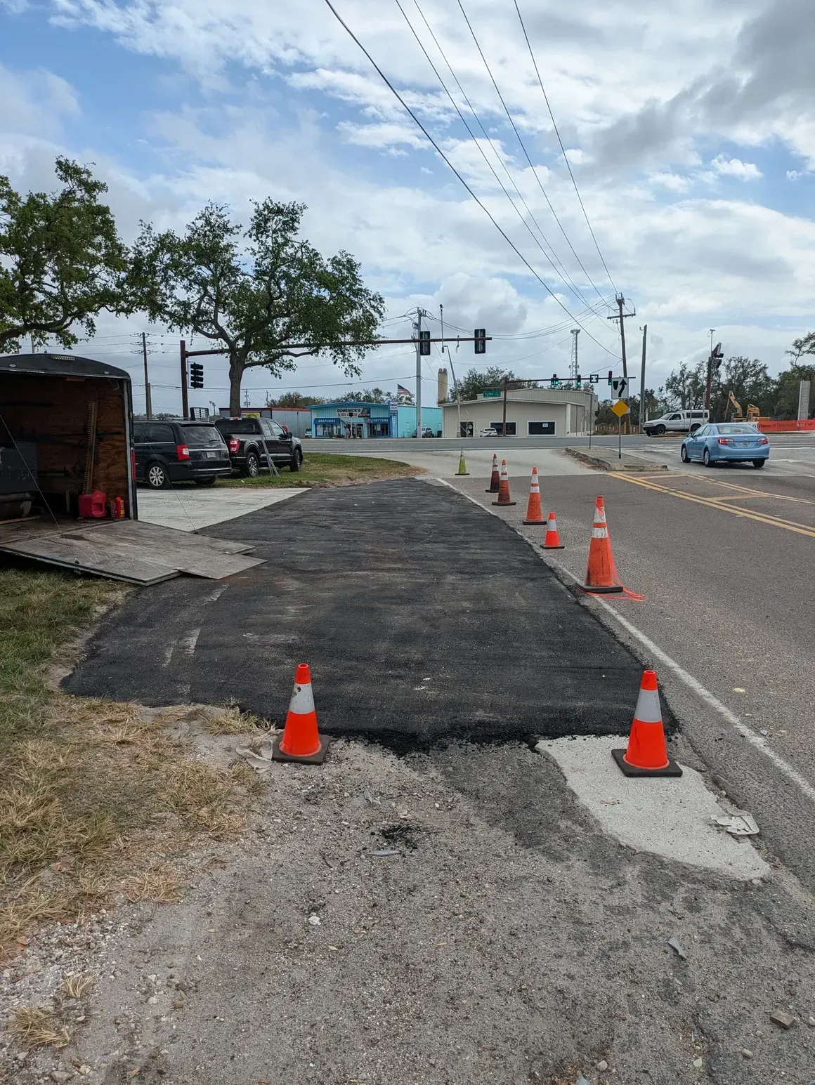 Orange cones mark a rough black-patched sidewalk beside a road under a cloudy sky.