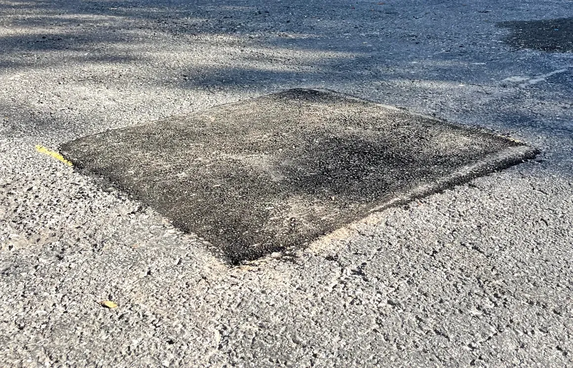 Pothole patch on a gravel road surface, dark asphalt square against lighter gravel.