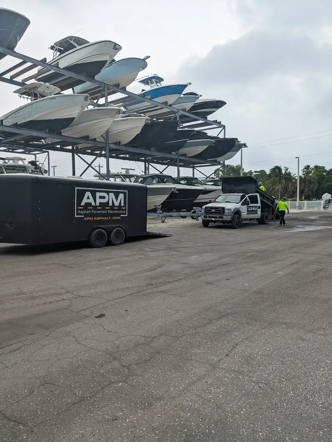 Stacked boats on trailers in a gravel lot beside an APM container truck and a white SUV.