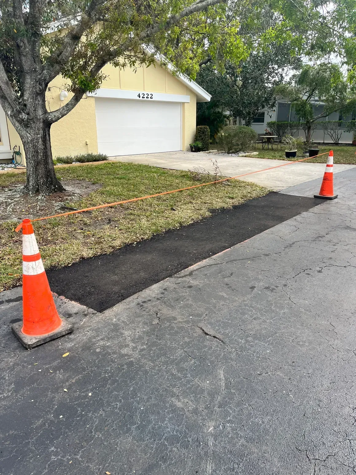 Freshly patched asphalt strip beside a driveway, bordered by two orange traffic cones near a garage.