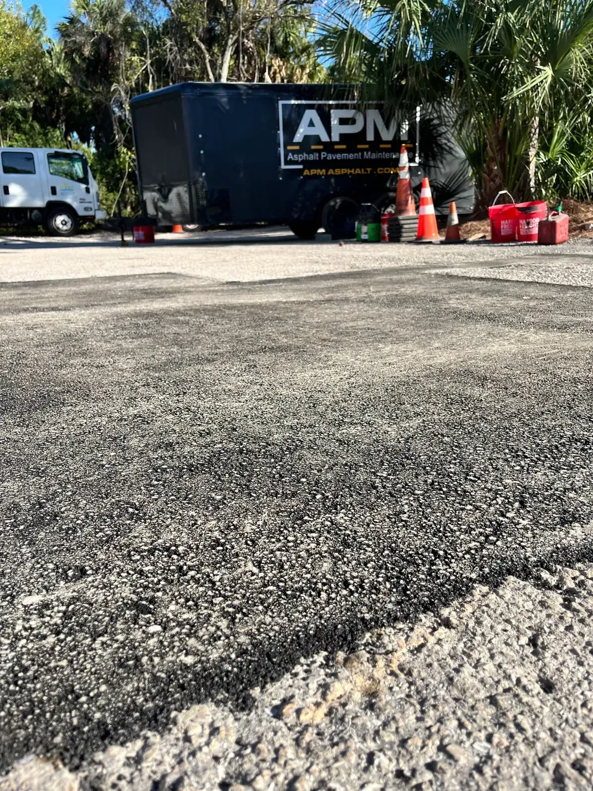 APM truck parked by traffic cones on a sunny roadside, with palm trees and rough pavement in foreground