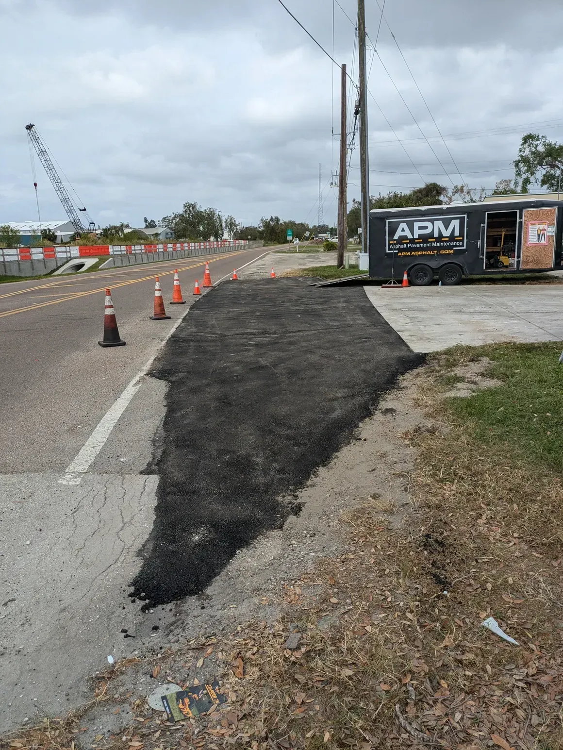 Roadwork beside a paved road with traffic cones and a fresh black asphalt patch near an APM sign