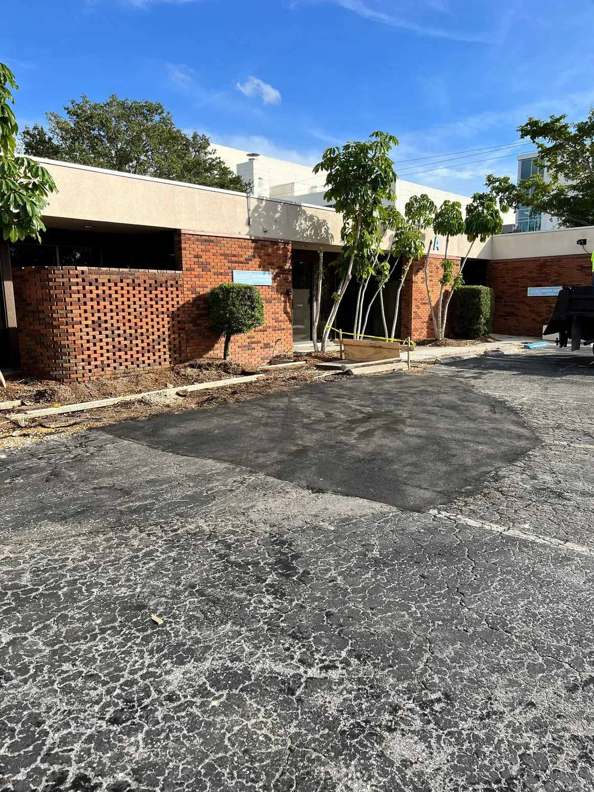 Brick building with black awnings and trees beside an empty asphalt parking lot under a blue sky