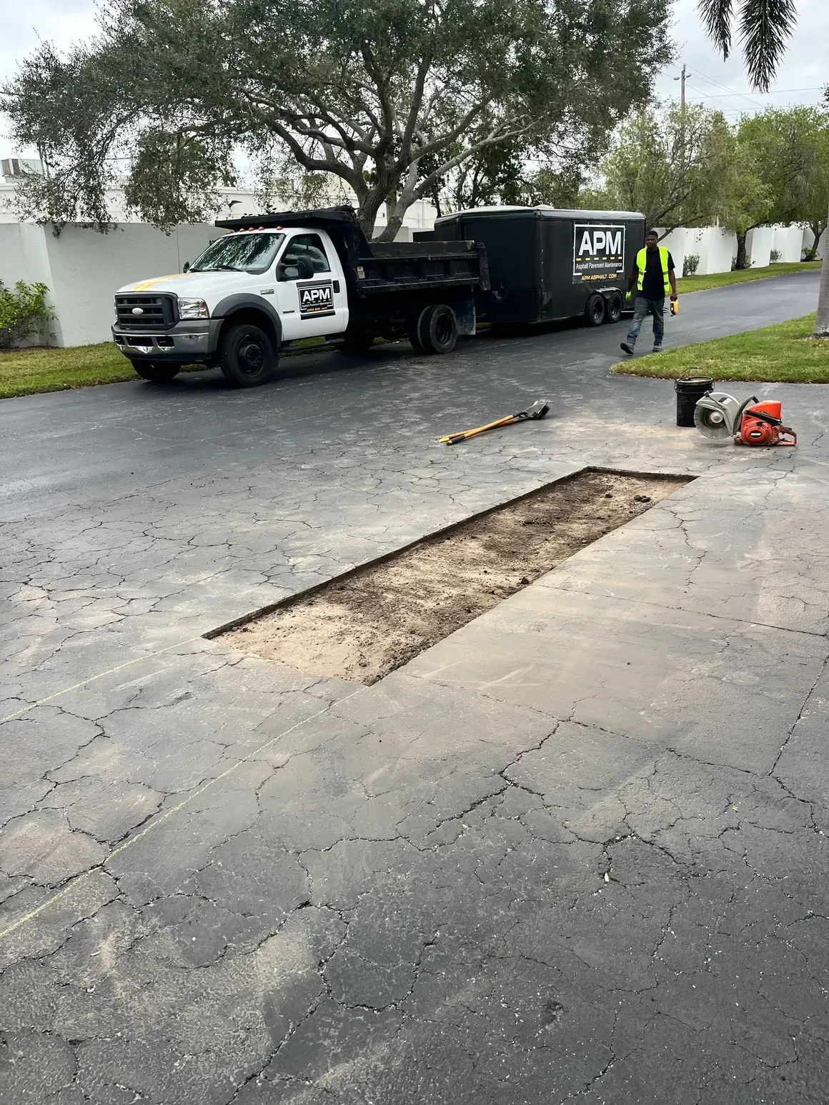 Road crew patching a pothole on a paved street beside a white truck and black trailer.