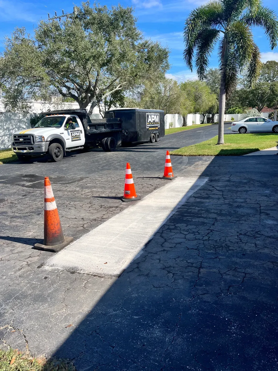 White truck and black trailer parked beside orange cones on a sunny roadside with palm trees.