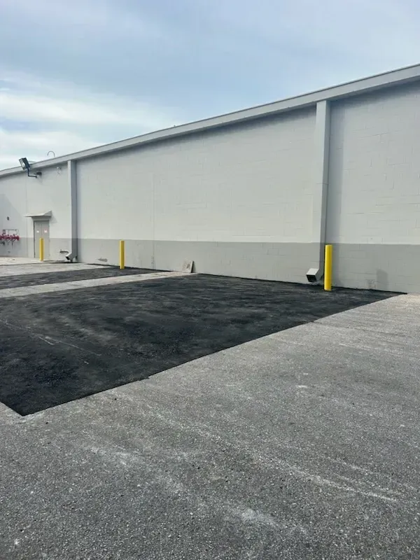 Empty industrial parking lot beside a gray warehouse wall with yellow bollards and dark asphalt patches