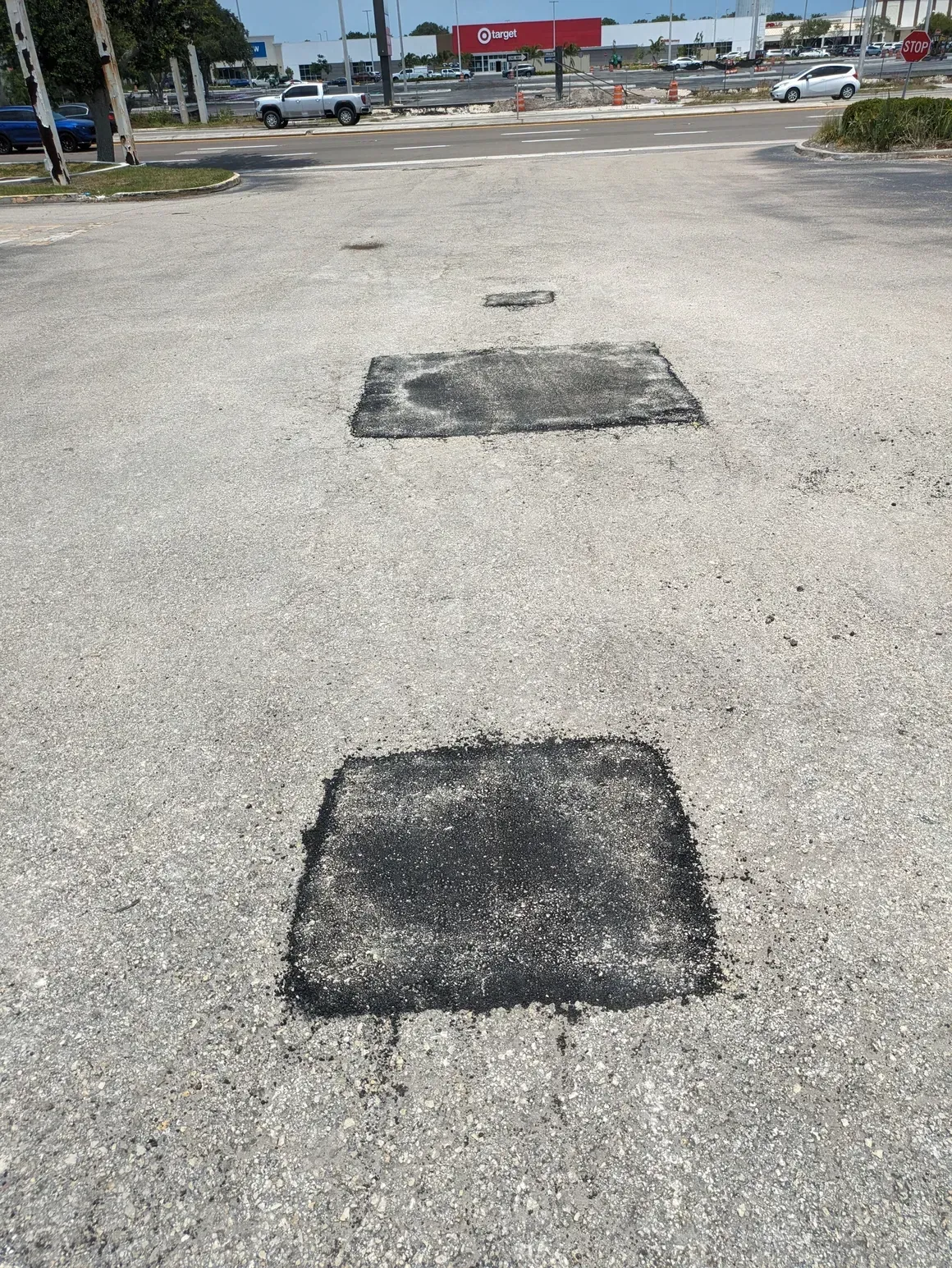 Parking lot with two faded square patches on cracked asphalt near a road and storefronts