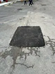 Large black asphalt patch on a cracked road surface, with two people in the background