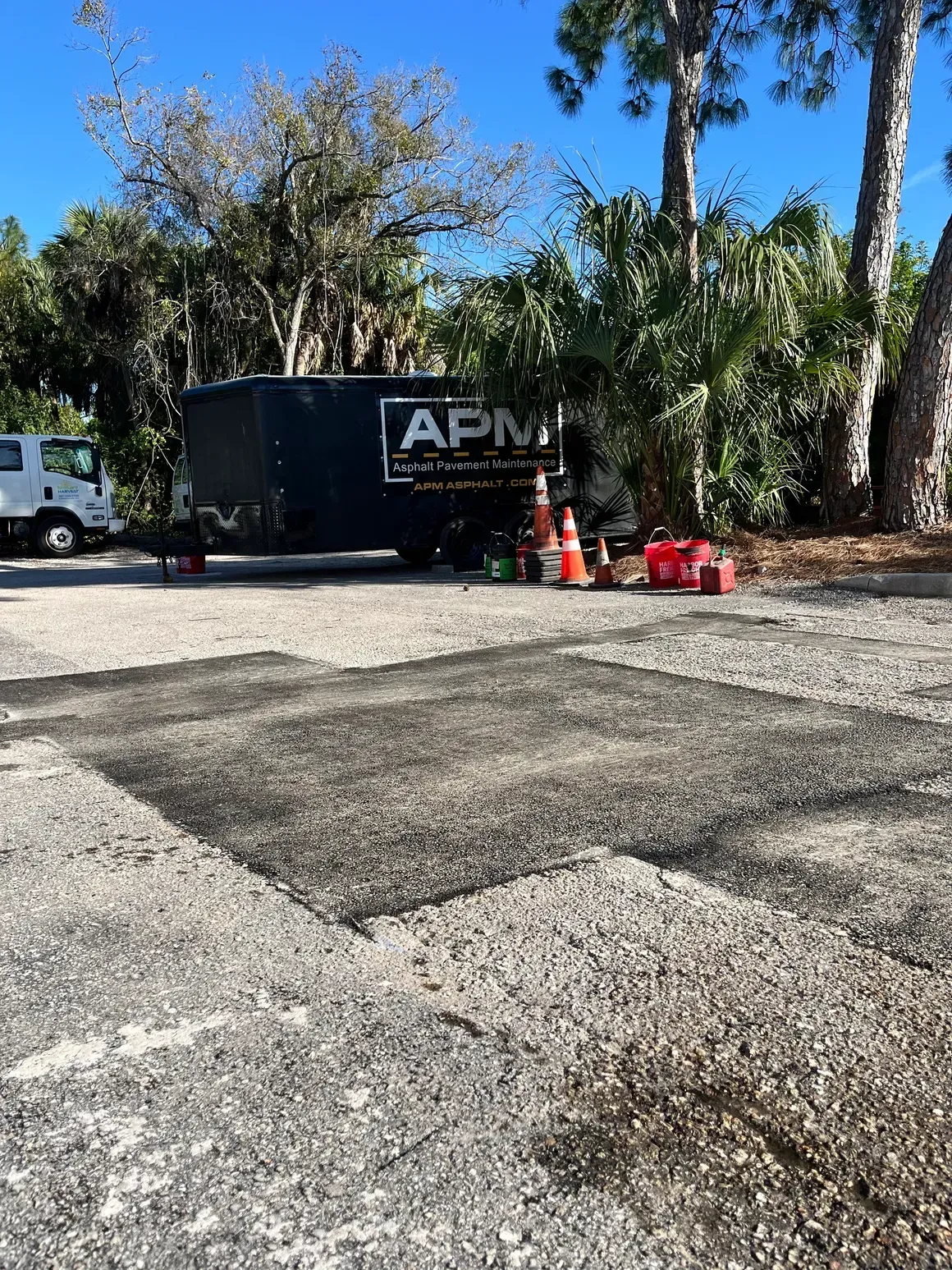 Parking lot with a black APM dumpster, orange cones, and trees under a blue sky.