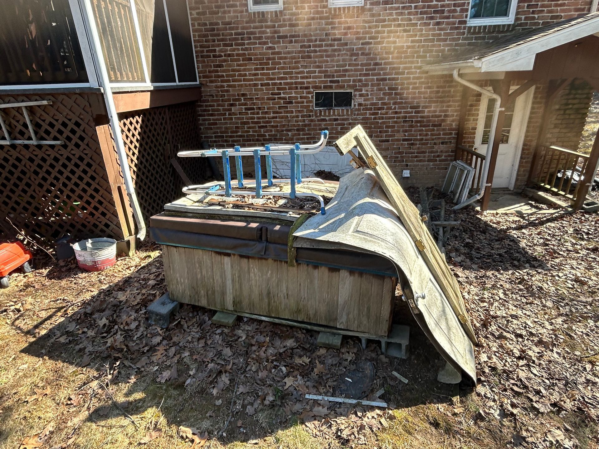 An old, damaged hot tub sits outdoors next to a brick building and a deck, covered in leaves.