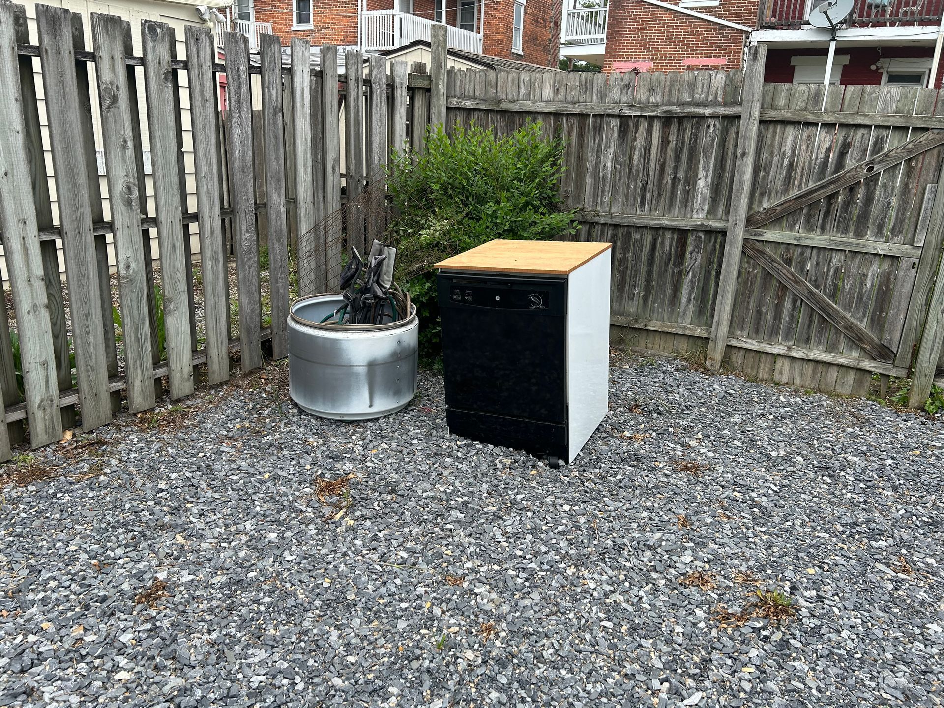 A dishwasher and metal drum sit in a gravel-covered yard, enclosed by a wooden fence.