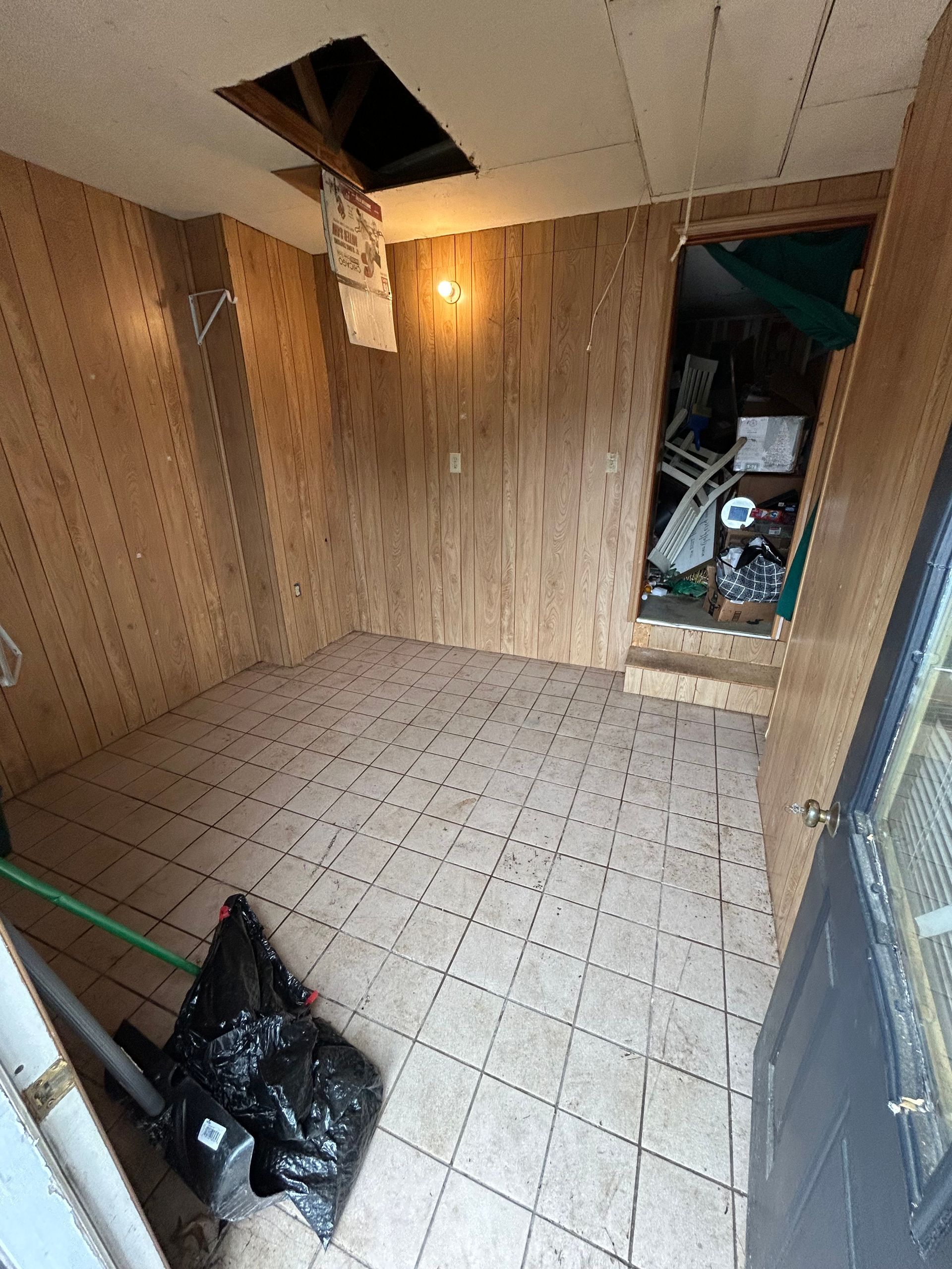 A small, cluttered room with damaged walls and a hole in the ceiling. Doorway leads to a cluttered space.