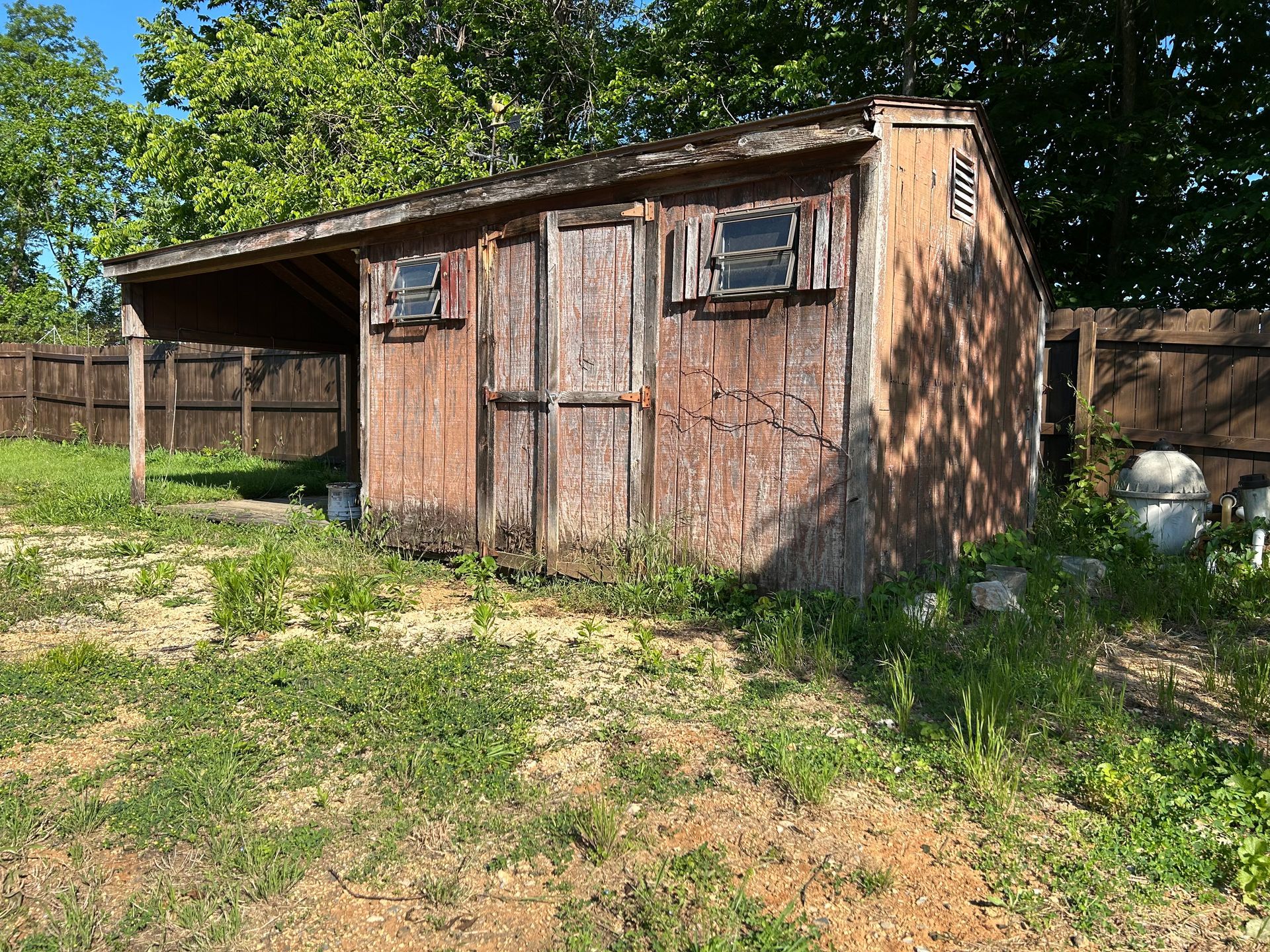 Weathered brown shed with a small overhang, surrounded by overgrown grass and a wooden fence.