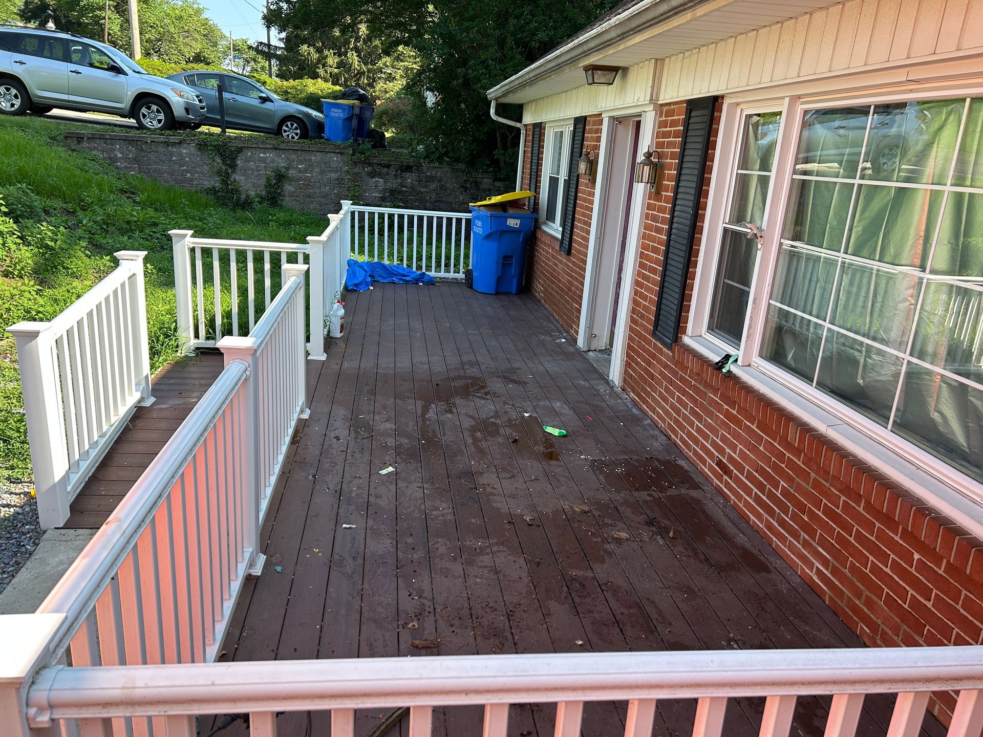 A wooden deck with white railings in front of a brick house; a blue trash bin sits on the deck.