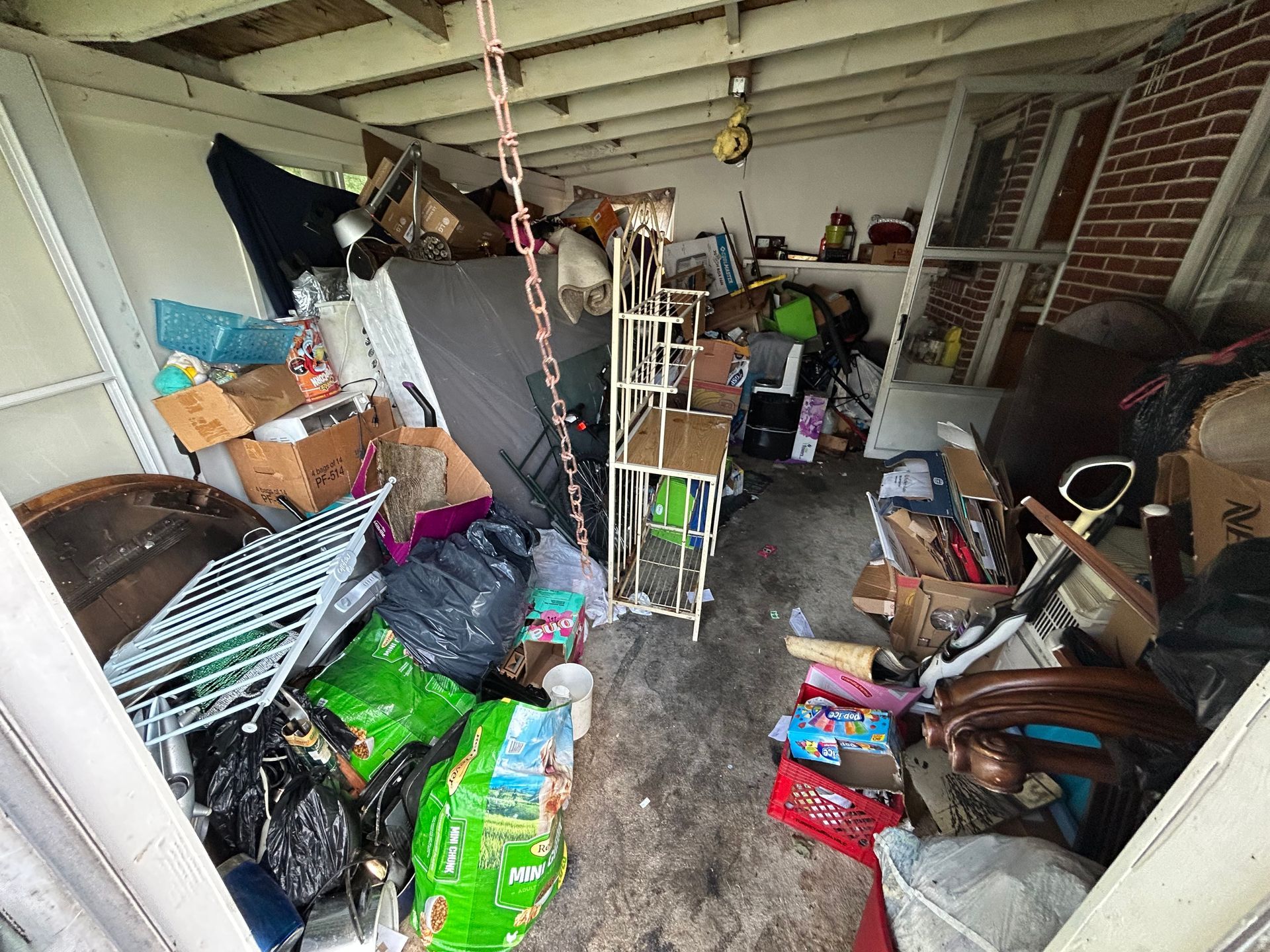 Cluttered shed interior with boxes, furniture, and various items piled haphazardly; chain hangs from ceiling.