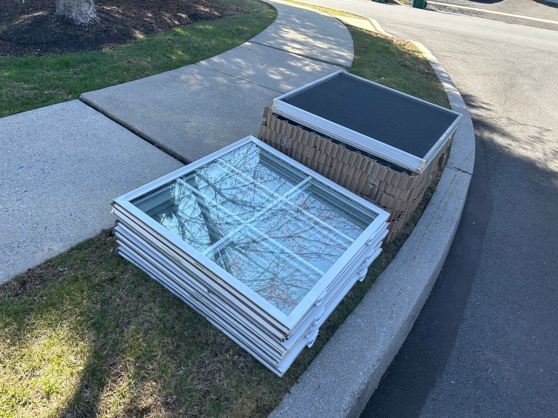 Stack of window panes and a solar panel on a curb, next to a sidewalk and grass.