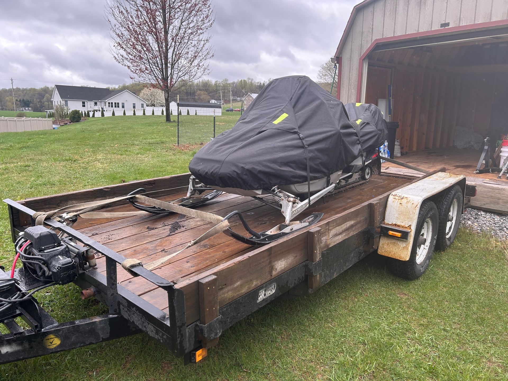 Snowmobile on a trailer near a garage, covered by a black tarp, green grass, cloudy sky.