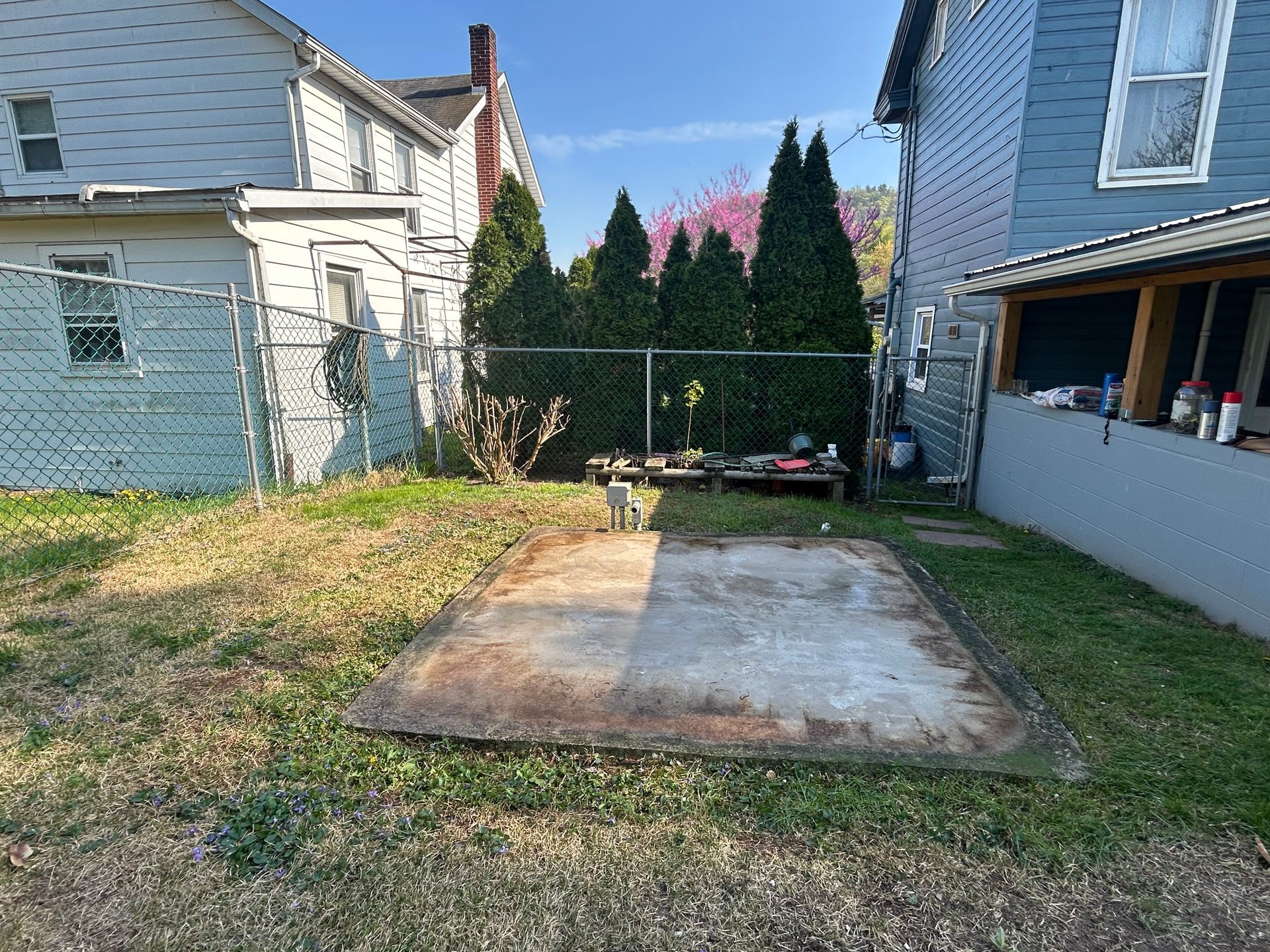Backyard with concrete slab, chain-link fence, small trees, and two-story houses in background.