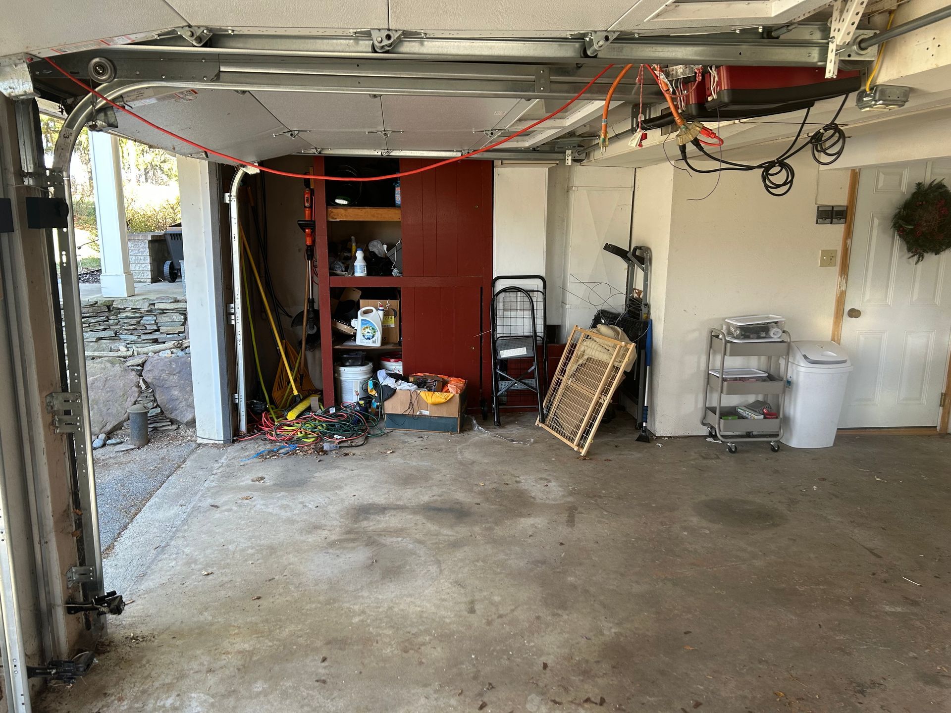 Garage interior with clutter, open door to outside. Gray concrete floor, red cabinet, tools.