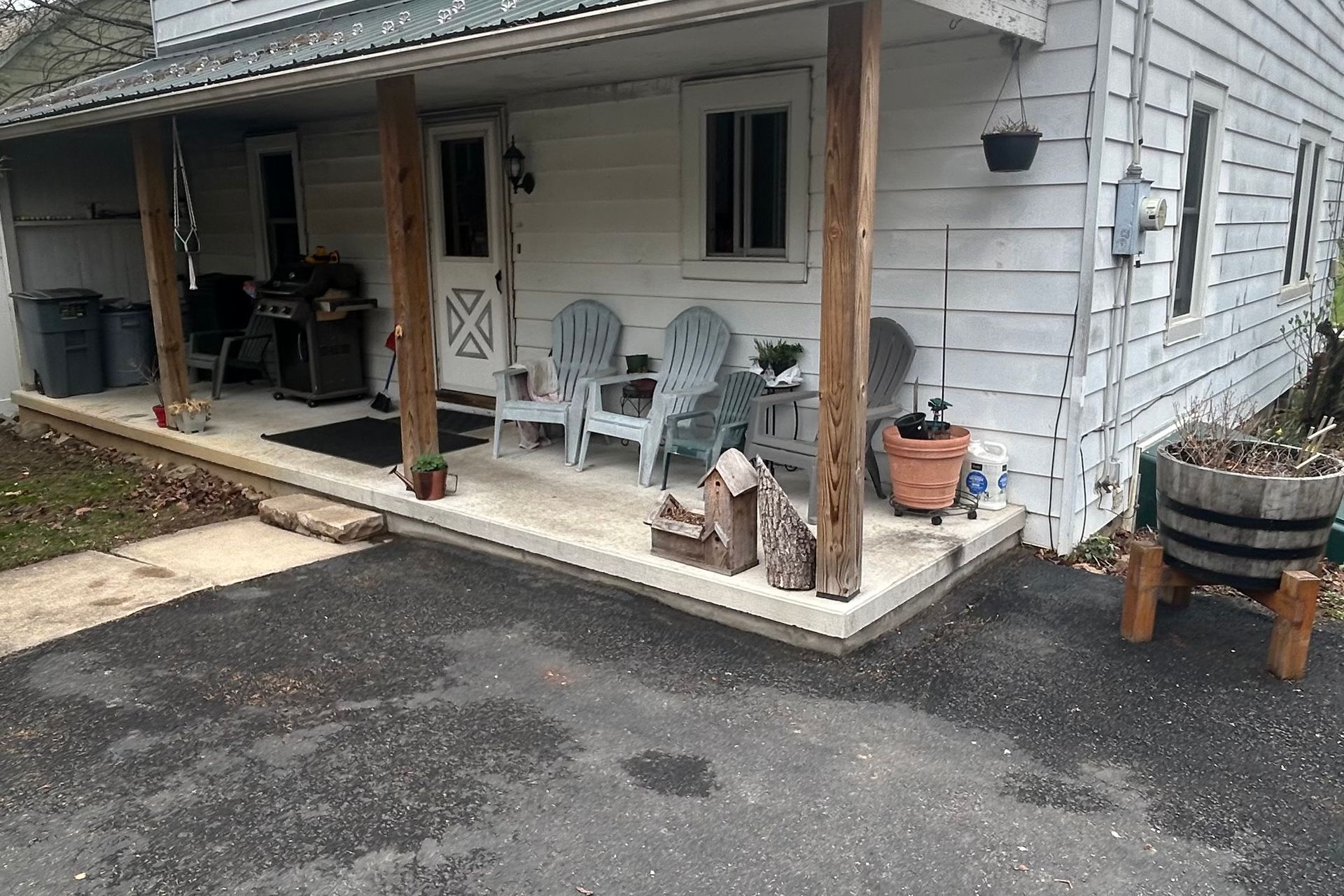 A white house with a covered porch, chairs, and a small planter sits next to a paved driveway.