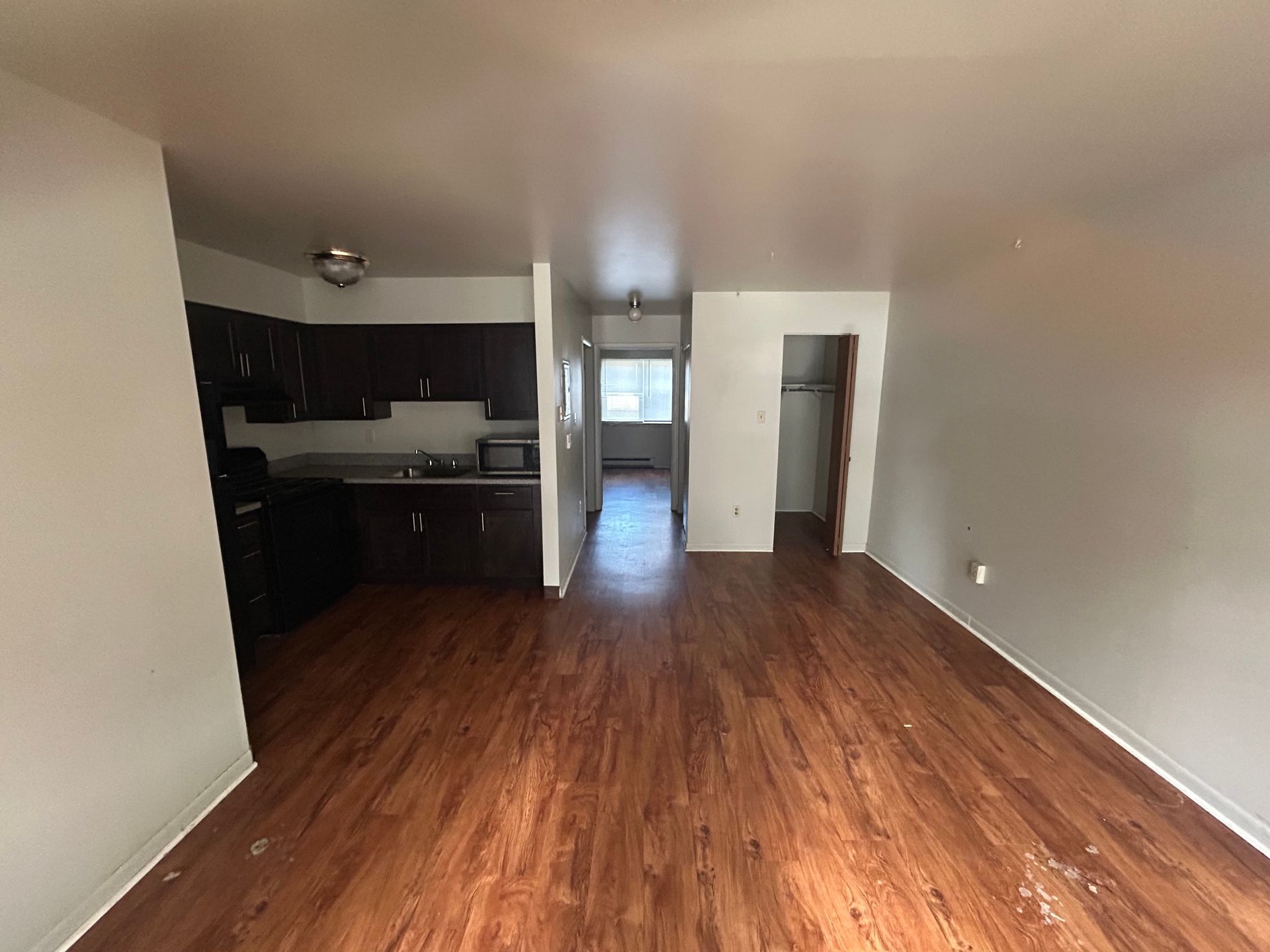 Interior view of a small apartment with dark brown cabinets, wood floors, and a narrow hallway with a window.