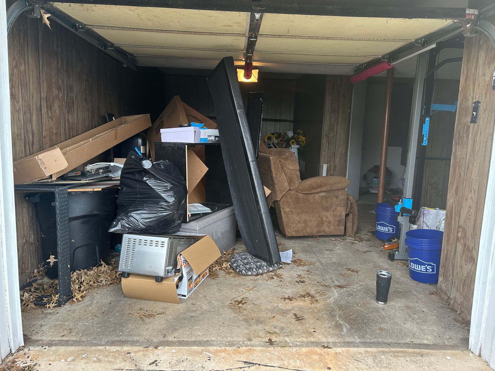 A cluttered garage with boxes, trash bags, and furniture; concrete floor.