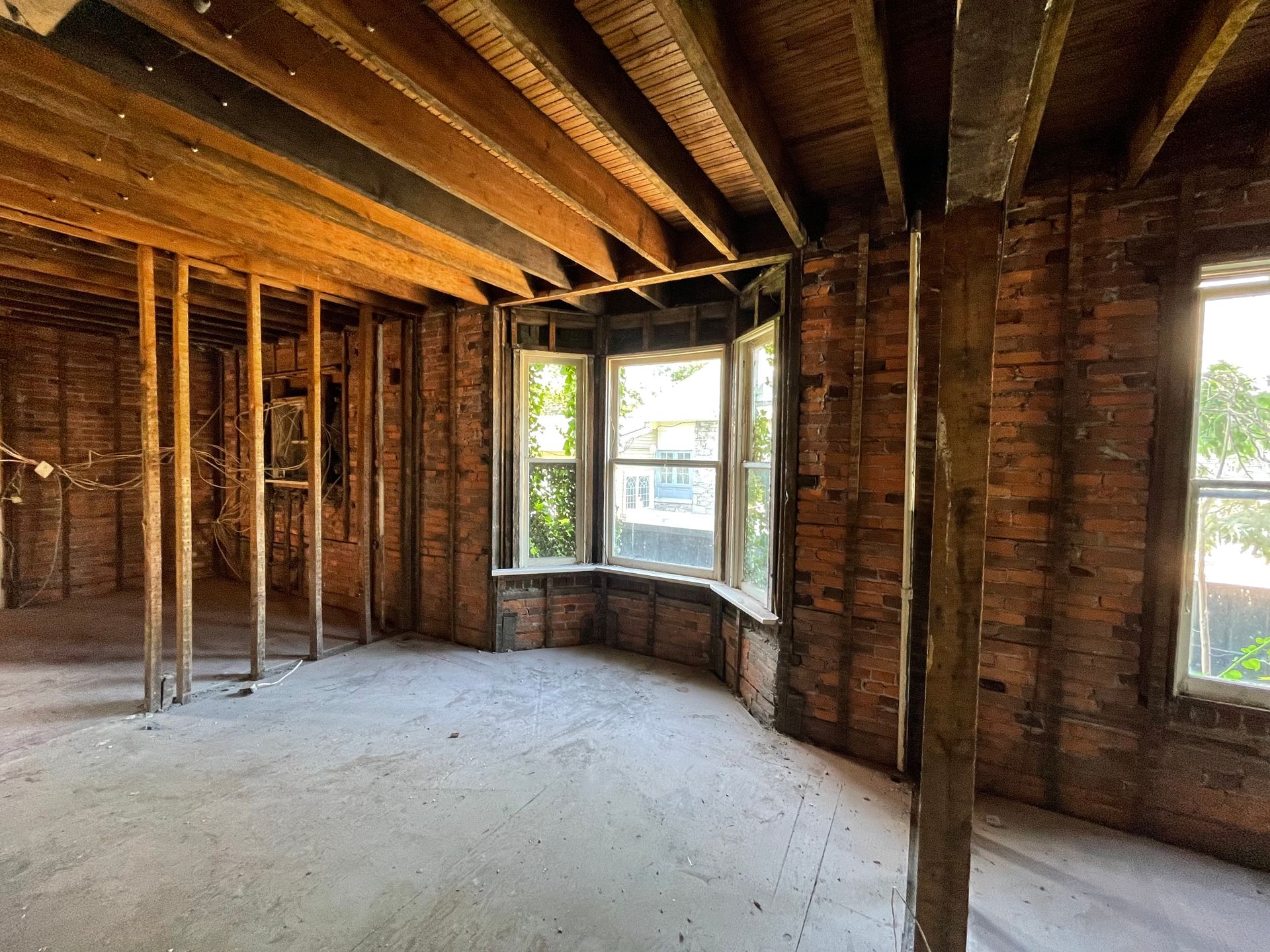 Room under renovation, exposed brick walls, bay window, wooden beams, studs, and a concrete floor.