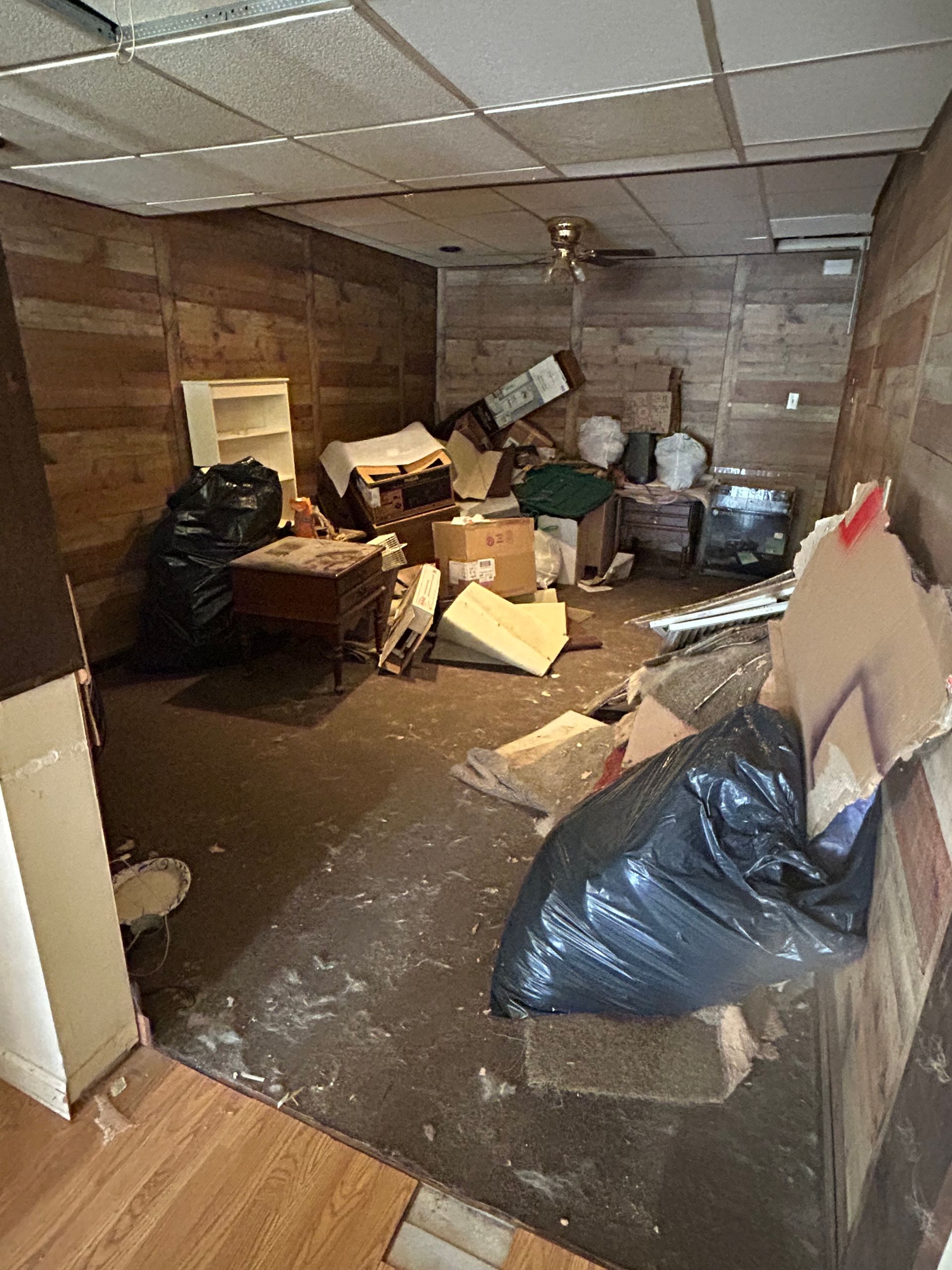 Cluttered room with trash bags, boxes, and furniture; wood panel walls and ceiling tiles.
