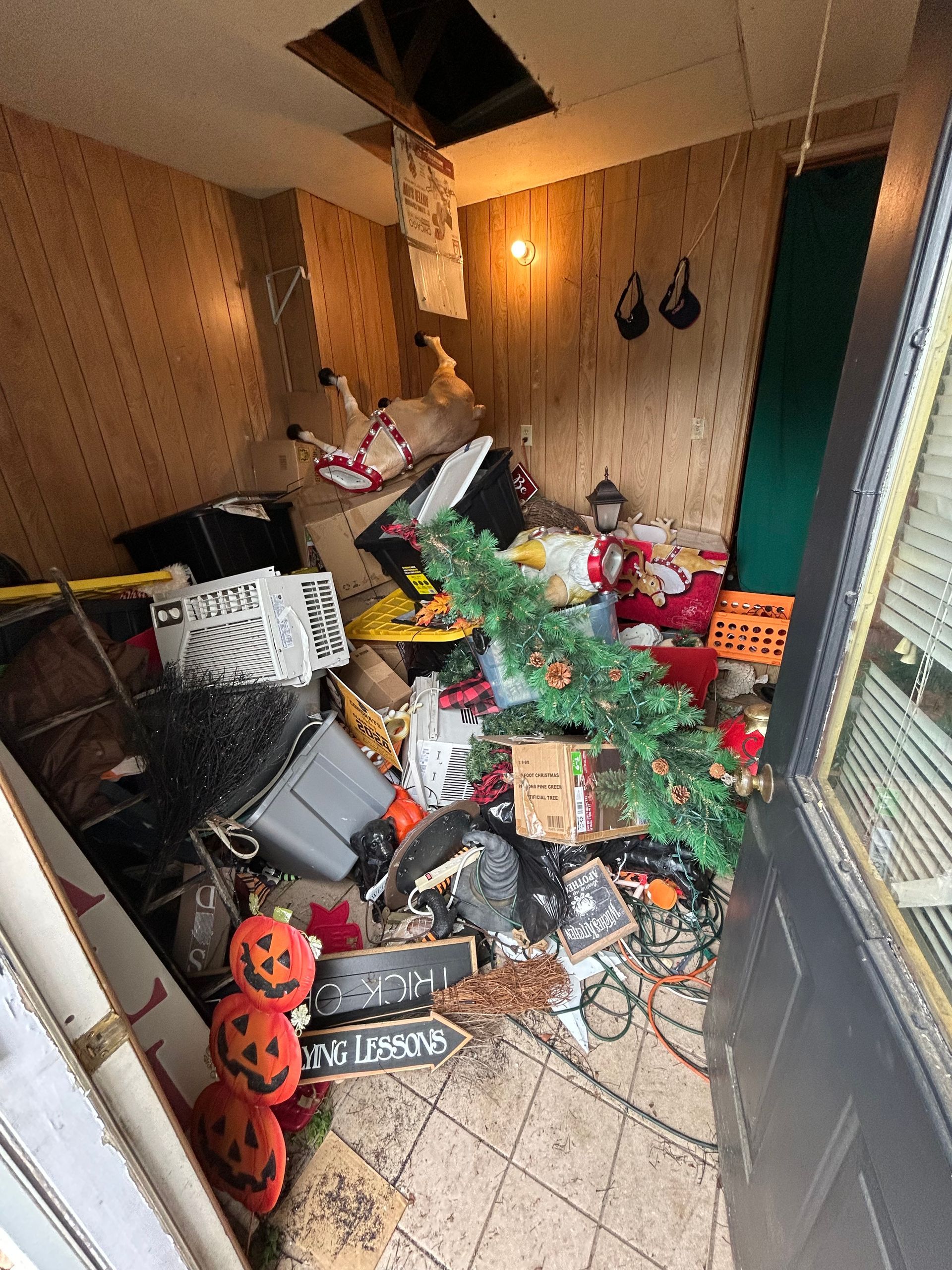 A cluttered room with a Christmas tree, Halloween decor, and a damaged ceiling.