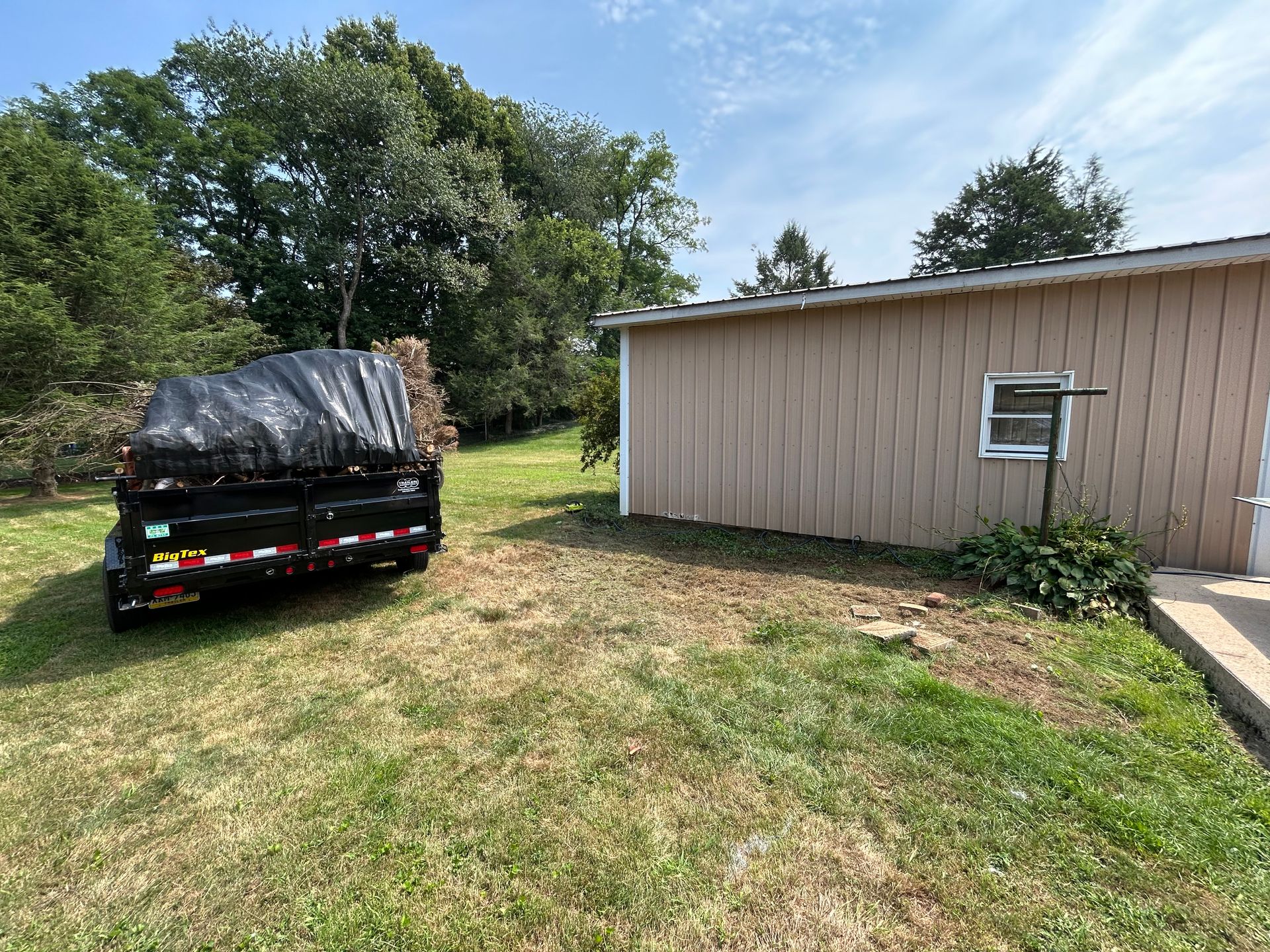 Trailer with tarp sits on grass beside a beige building under a blue sky.