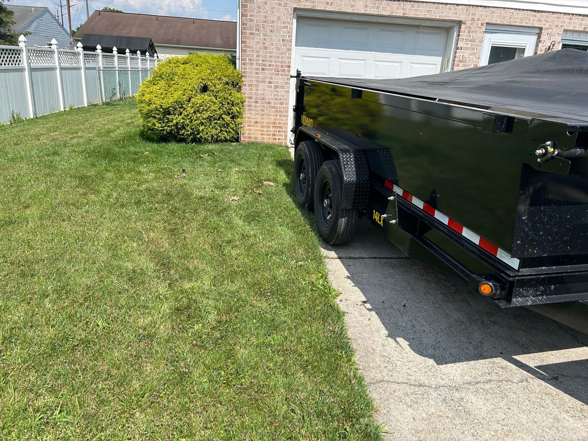 Black trailer partially in garage next to grassy yard, white fence, and a shrub.