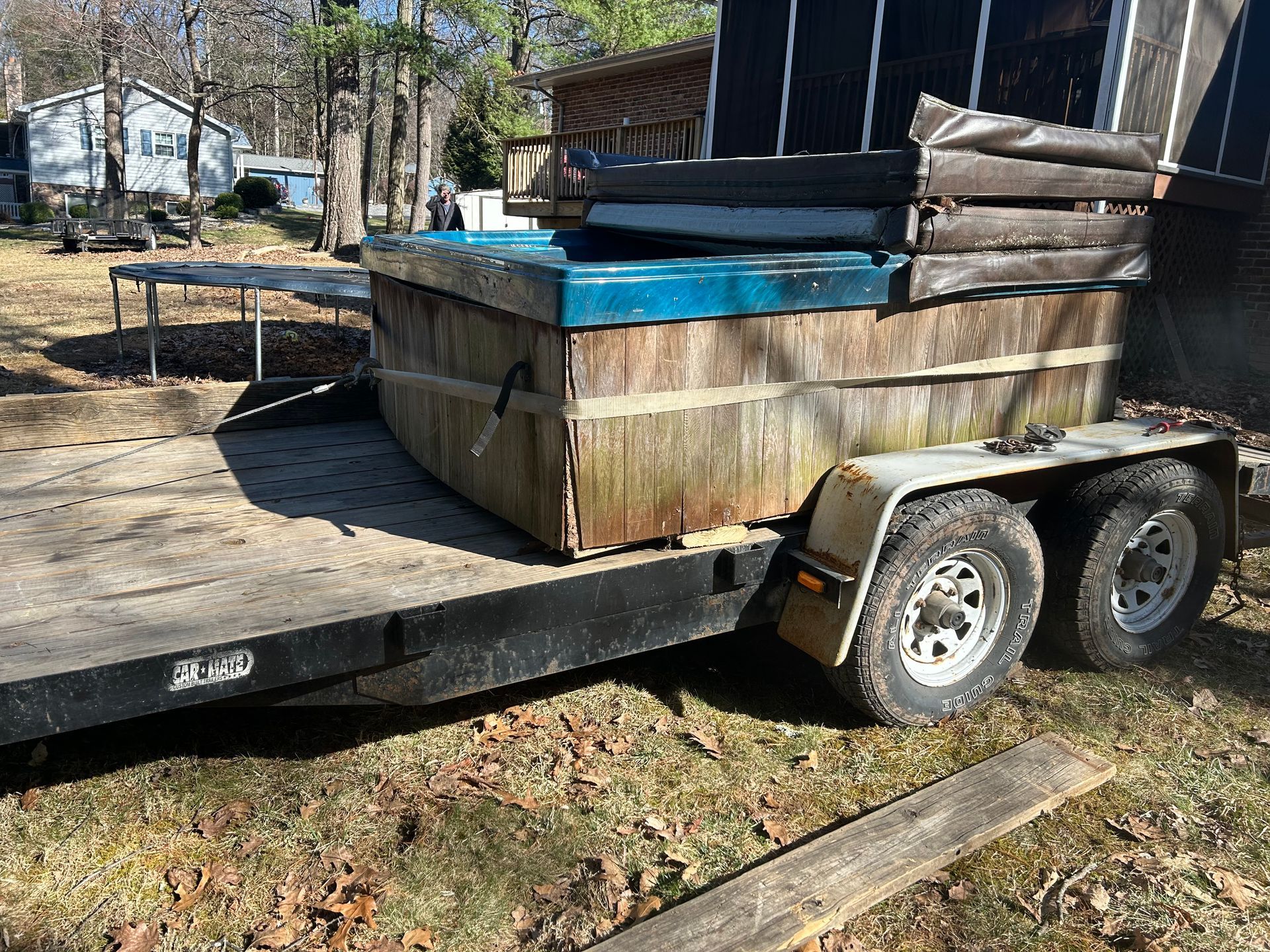 A weathered hot tub on a trailer, outdoors. Brown wooden sides, blue interior, and two tires visible.