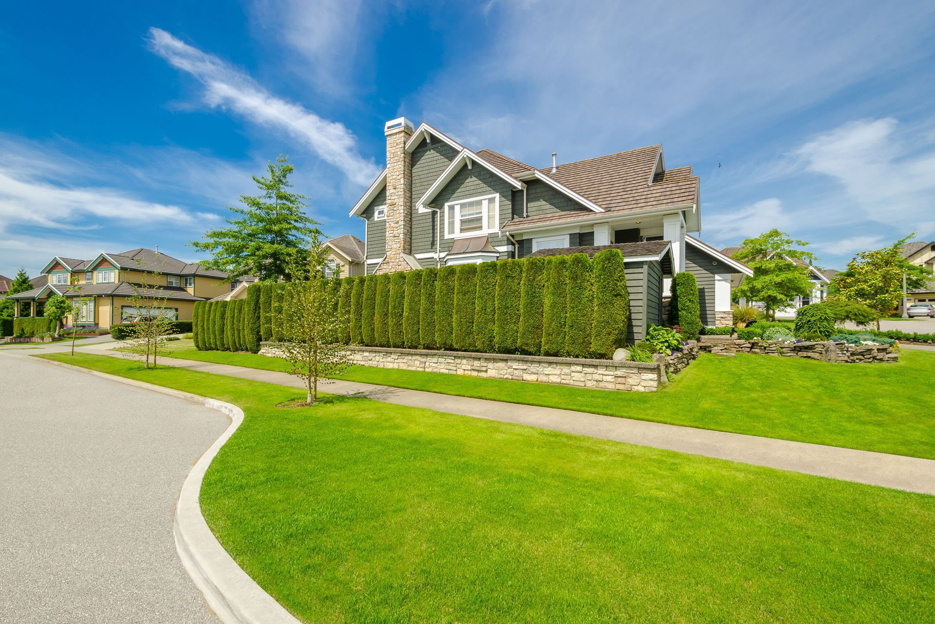 A suburban two-story house with dark siding and a stone fence, surrounded by green lawns under a bright blue sky.