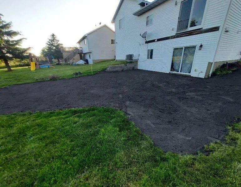 A white house exterior with a freshly leveled dark soil backyard area adjacent to a green grass lawn.