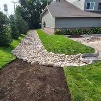 A rock-lined drainage swale curves alongside a patch of green lawn next to a house, with bare soil in the foreground.