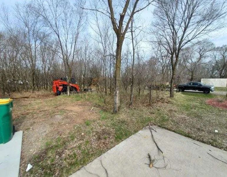 A red skid steer clears brush in a wooded yard near a concrete driveway and a black pickup truck.