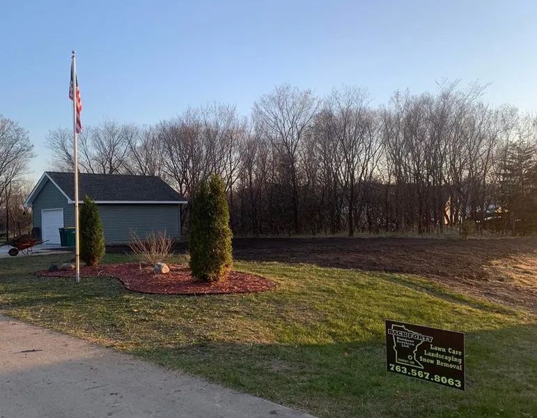 A flag pole, a garage, and a landscaped bed with two evergreens stand near a lawn with a landscaping business sign.