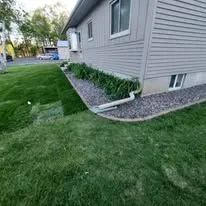 A corner of a grey-sided house with a white downspout, bordered by a gravel garden bed and newly laid green sod.