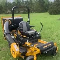 A yellow Hustler zero-turn lawn mower with a large black grass collection chute attached, parked on a grassy lawn.