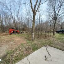 A red tractor works in a wooded lot near a paved driveway, with a black truck visible in the background.