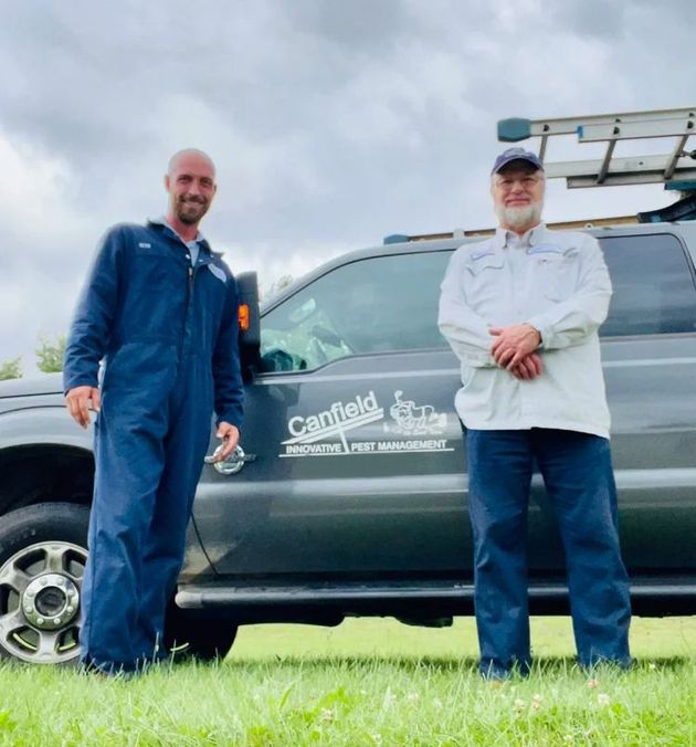 Two workers in blue uniforms standing beside a dark work truck on grass, one holding a tool rack.