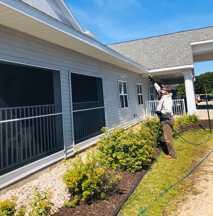 Person pressure-washing a gray house exterior beside screened porches and shrubs.