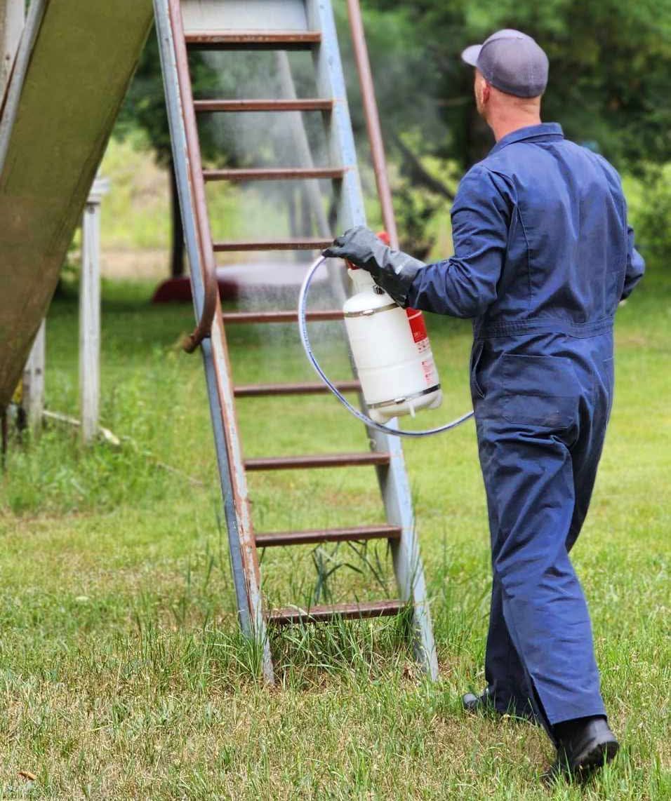 Worker in blue coveralls carrying a backpack sprayer beside a metal ladder outdoors