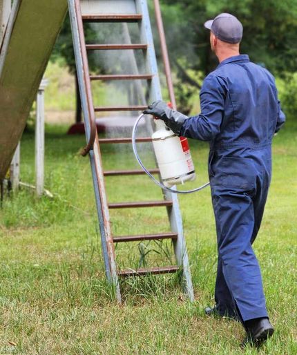 Worker in blue coveralls carrying a backpack sprayer beside a metal ladder outdoors