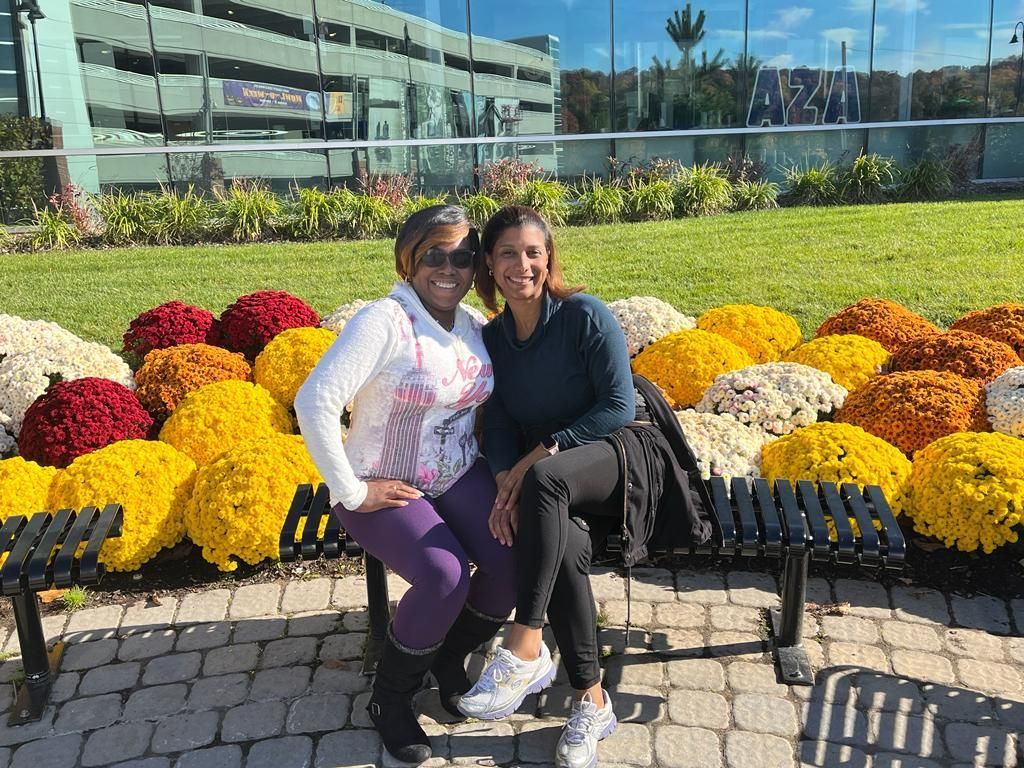 Two people seated on a bench, smiling, next to colorful flowers. Building in background. Sunny day.