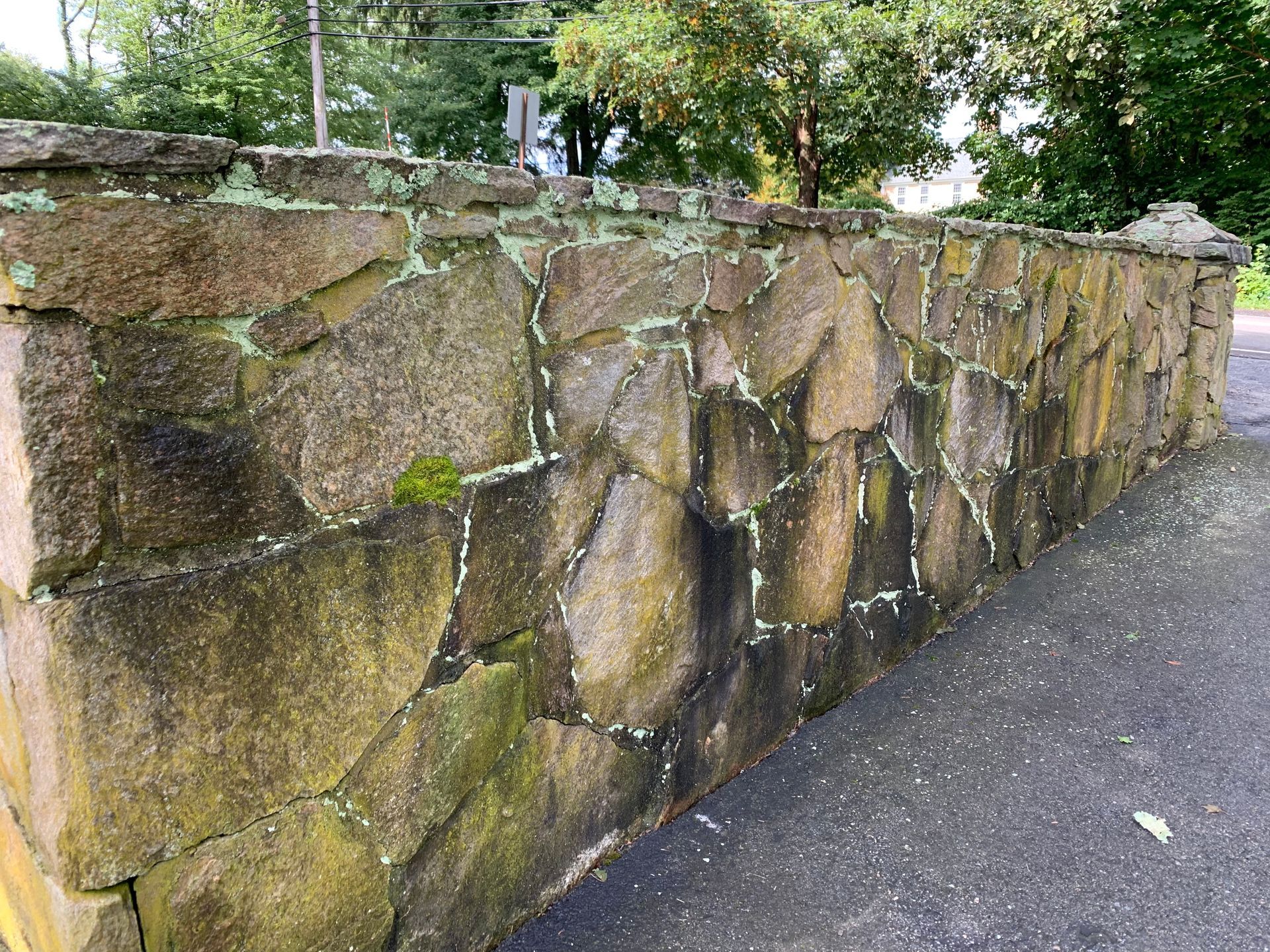 A large stone wall with moss growing on it is next to a road.