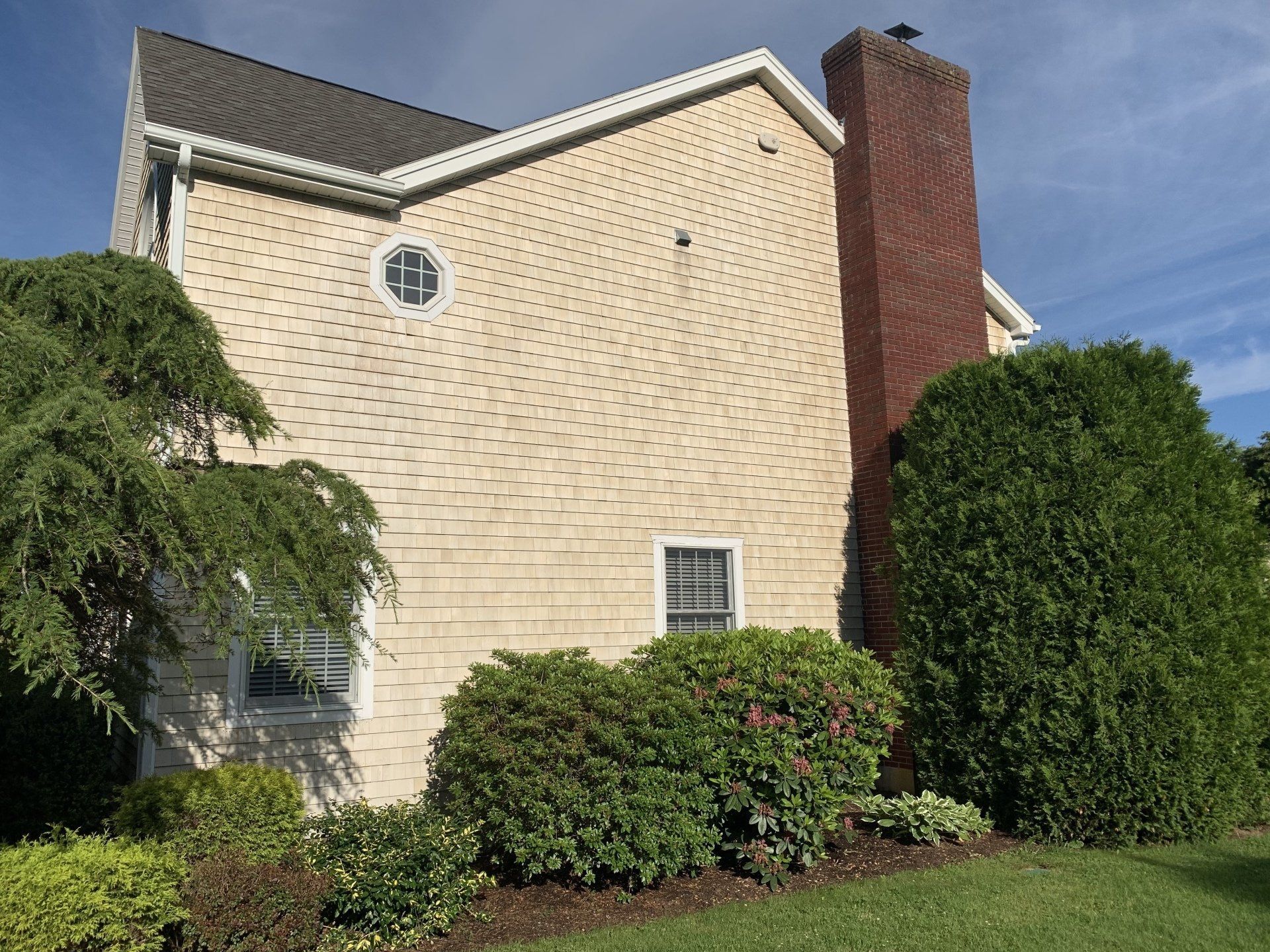 A large house with a brick chimney is surrounded by bushes and trees.